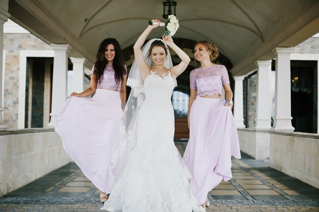 Preciosa novia y damas de honor en vestidos violetas y rosas de pie bajo los arcos