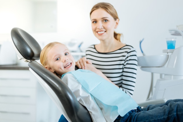 Precioso apoyo. encantadora niña y su madre posando para la cámara mientras la niña sentada en la silla del dentista y su madre