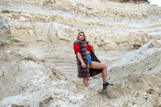 Precioso modelo de moda usar abrigo de piel de pie en la cantera de arena vacía y seca en un cálido día soleado de otoño