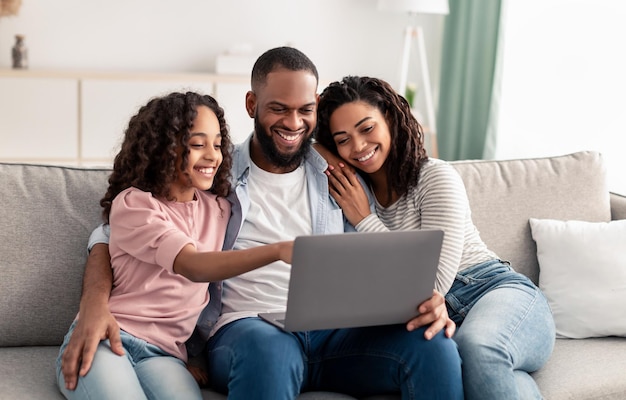 Precioso tiempo con la familia. Retrato de felices padres afro y una niña linda usando una computadora portátil juntos, viendo fotos o películas, navegando por Internet, eligiendo regalos en línea sentados en el sofá de la sala de estar