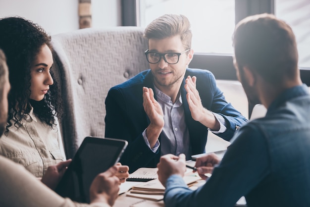 Precisamos de mudanças! Jovem bonito de óculos gesticulando e discutindo algo com seus colegas de trabalho enquanto está sentado à mesa do escritório