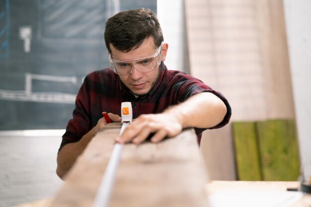 Precisión a lo largo de un joven carpintero serio que trabaja con madera en su taller