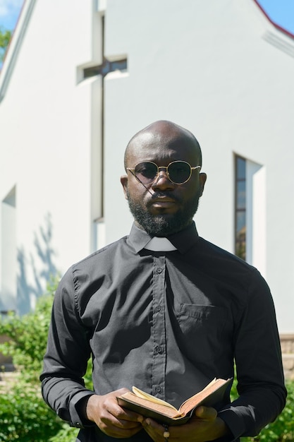 Predicador afroamericano en camisa negra con cuello clerical sosteniendo biblia