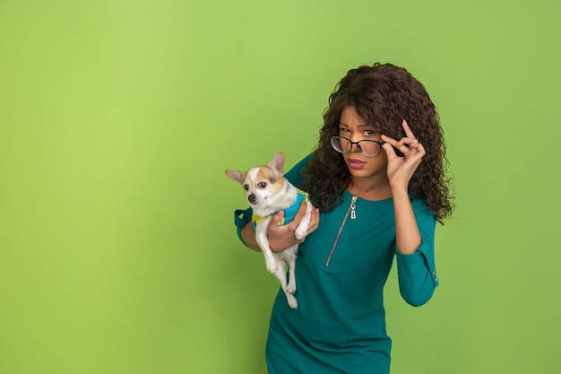 Preguntando, dudas. Retrato de mujer joven afroamericana en gafas aislado sobre fondo verde de estudio. Preciosa modelo con perrito. Concepto de emociones humanas, expresión facial, ventas, publicidad.