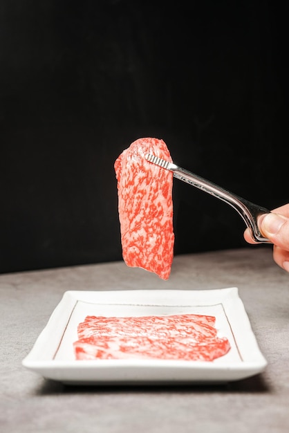 Premium Rare Slices Sirloin Wagyu A5-Rindfleisch mit hochmarmorierter Textur auf einer Keramikplatte, die von einer Grillzange mit der Hand aufgenommen wird. Serviert für Yakiniku Sukiyaki und Shabu Image mit Kopierbereich