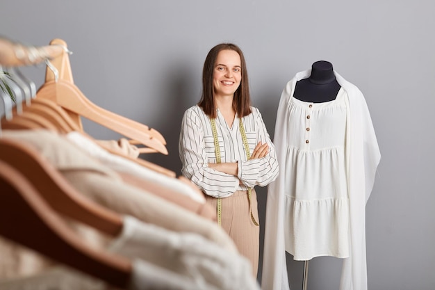 Prendas únicas clientes felices Atractiva mujer sonriente sastre de pie cerca de un vestido nuevo en maniquí aislado sobre fondo gris mantiene las manos cruzadas mirando a la cámara