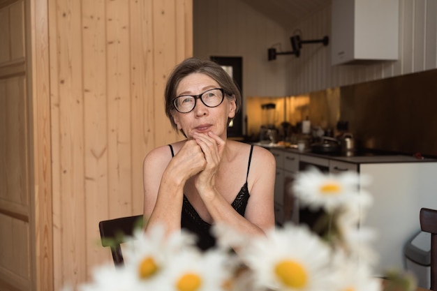 Preocupada anciana de 60 años con gafas sentada en la mesa de la cocina