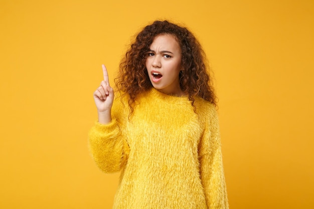 Preocupada joven afroamericana irritada con suéter de piel posando aislada en un fondo amarillo anaranjado en el estudio. Concepto de estilo de vida de las personas. Simulacros de espacio de copia. Jurando, apuntando con el dedo índice hacia arriba.