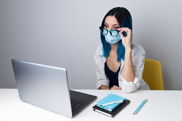 Preocupada mujer con cabello azul en máscara protectora mirando en contenido portátil sentado en la oficina en casa