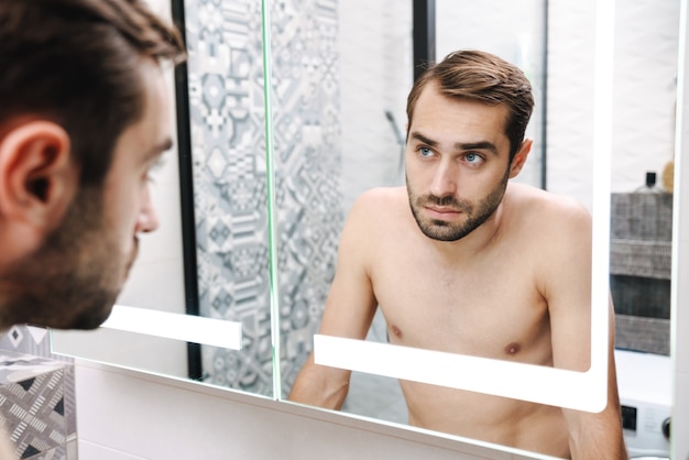Preocupado joven descamisado mirando a sí mismo en el espejo del baño