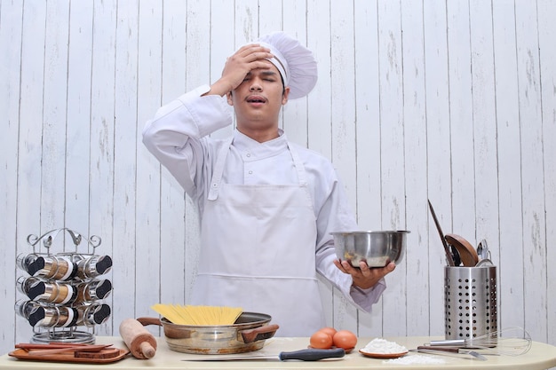 Preocupado por el trabajo del joven chef en la mesa, los utensilios de cocina se ponen la mano en la cara por una expresión decepcionada, fa épica