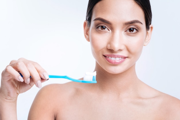 Preocuparse por una bonita sonrisa. Agradable joven alegre con hombros desnudos sosteniendo un cepillo de dientes con un poco de pasta de dientes y sonriendo