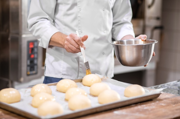 Preparação para assar. Mãos masculinas com tigela e escova especial cobrindo pãezinhos na bandeja com esmalte de confeitaria