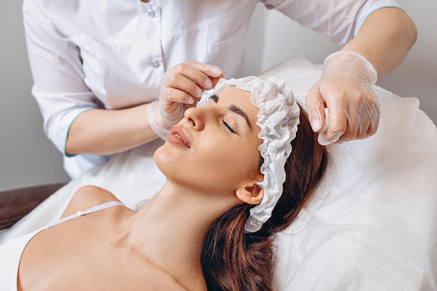 Preparación antes de iniciar el procedimiento en el salón de belleza. Un trabajador de un salón de belleza con uniforme blanco y guantes protectores le pone una gorra protectora a un cliente joven. Seguridad antes de prestar servicios.