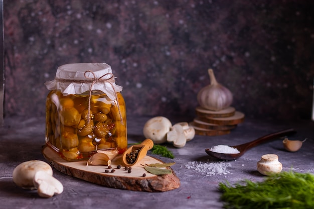 Preparación de champiñones enlatados fermentados con laurel, pimienta, sal, ajo y eneldo en un frasco de vidrio sobre un soporte de madera en un tono bajo