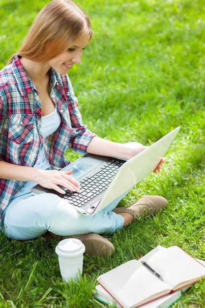 Preparación para exámenes al aire libre. Hermosa joven estudiante trabajando en la computadora portátil y sonriendo mientras está sentado en un parque con libros a su alrededor