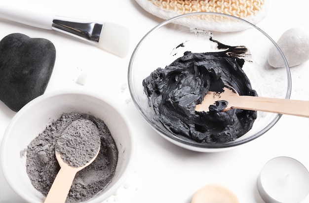 Preparación de mascarilla de barro negro cosmética casera en un tazón de vidrio sobre fondo blanco.