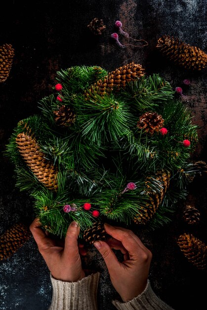 Preparación para vacaciones de navidad. Mujer que adorna la guirnalda verde de Navidad con piñas y bayas rojas de invierno, en copyspace oxidado oscuro, vista superior, manos femeninas