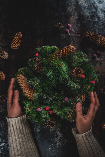 Preparación para vacaciones de navidad. Mujer que adorna la guirnalda verde de Navidad con piñas y bayas rojas de invierno, en copyspace oxidado oscuro, vista superior, manos femeninas