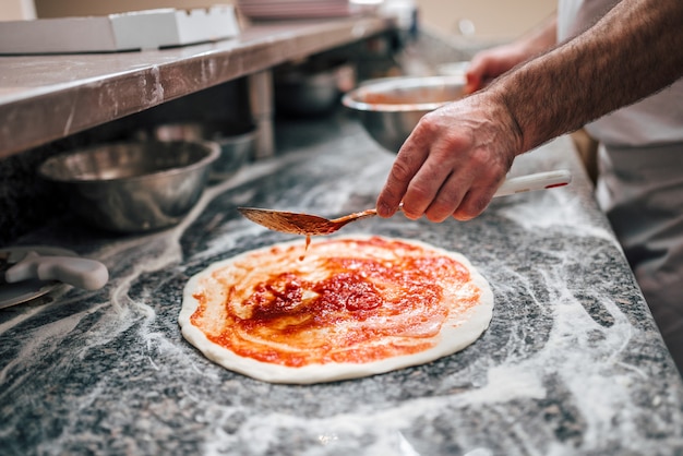 Preparando pizza. Mano del chef añadiendo salsa de tomate a la masa de pizza.