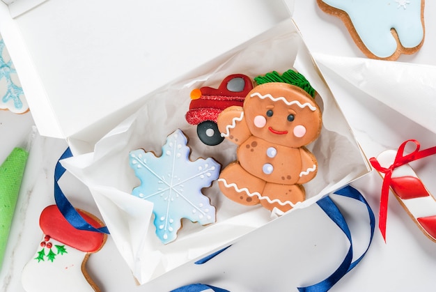 Preparando-se para o Natal, decorando o tradicional pão de gengibre com cobertura de açúcar multicolorido, biscoitos, pão de mel em uma caixa de presente branca, com laço de fita, em uma mesa de mármore branco vista superior copyspace