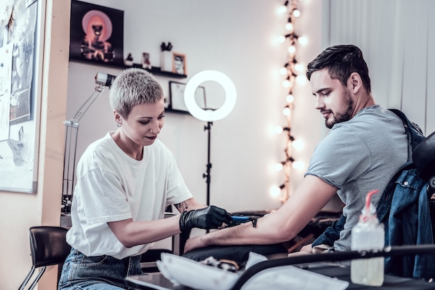 Preparando-se para o trabalho. O extraordinário mestre da tatuagem de cabelo curto raspando o cabelo da mão do cliente tornando-o suave