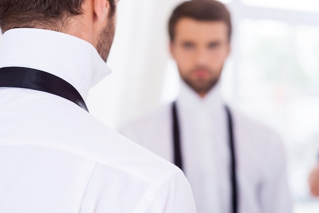 Preparándose para un día especial. Vista trasera del joven con camisa blanca y corbata desatada de pie contra el espejo