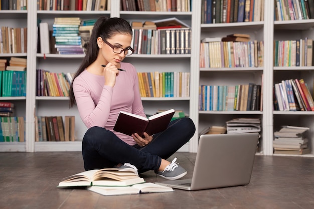 Preparándose para los exámenes. Mujer joven pensativa sosteniendo un libro y mirando la computadora portátil mientras está sentado en el suelo en la biblioteca