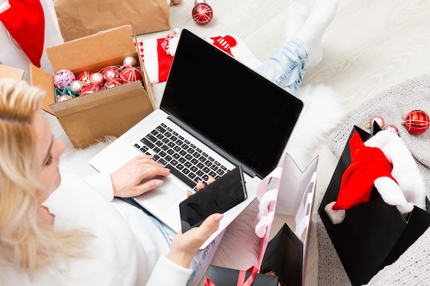 Preparándose para la fiesta de Navidad. Mujer ordenando regalos y decoraciones en una computadora portátil, sentada entre cajas y paquetes de regalos, espacio de copia