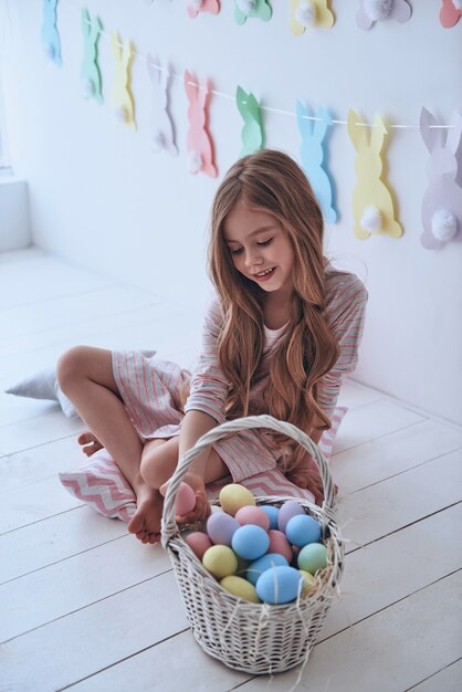 Preparándose para la Pascua. Hermosa niña sosteniendo un huevo de Pascua y sonriendo mientras está sentado en la almohada con decoración en el fondo