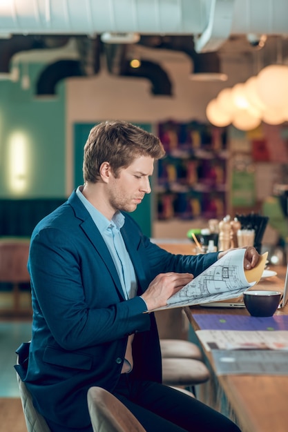 Preparándose para la reunión. Hombre de negocios joven serio mirando el plan de construcción en la carpeta sentado con un café en la cafetería esperando una cita