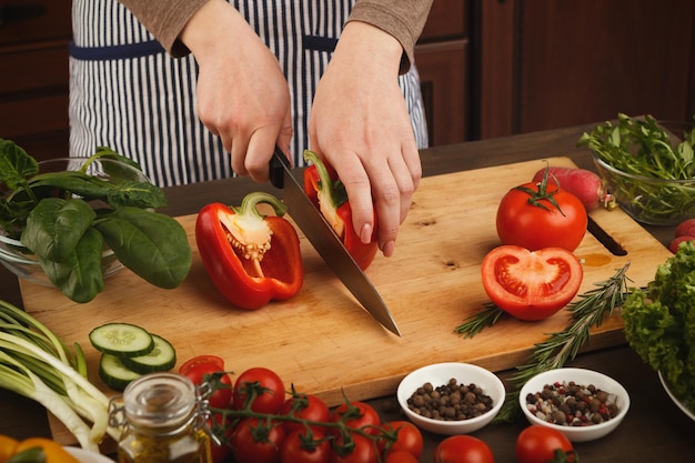 Preparar ensalada de verduras frescas en la mesa de la cocina, vista lateral. Cortar pimiento rojo maduro sobre tabla de madera, espacio de copia