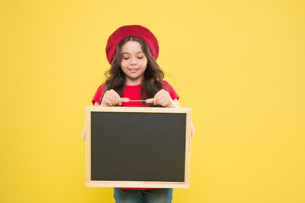 Presentación del producto niña pequeña con espacio de copia en el tablero de la escuela tablero de publicidad de regreso a la escuela para promoción ventas de compras escolares niño sobre fondo amarillo niña feliz en boina francesa