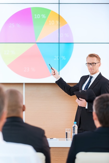 Foto presentación en sala de conferencias. hombre joven confiado en ropa formal que señala la pantalla de proyección con un gráfico mientras hace una presentación en la sala de conferencias con personas en primer plano