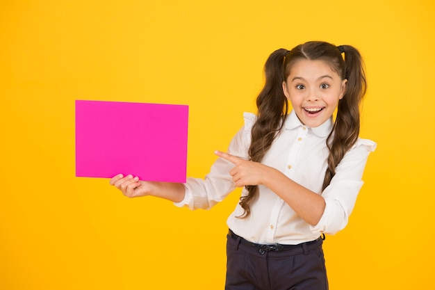 Presentación de su producto Niña feliz señalando con el dedo el papel rosa en blanco para publicidad de productos sobre fondo amarillo Producto de regreso a la escuela Llamando la atención sobre el espacio de copia del producto o evento