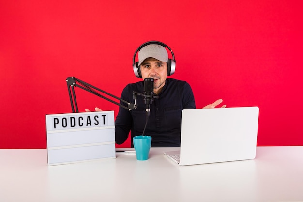 Presentador de hombre con gorra en estudio de grabación de radio podcast haciendo gestos con las manos, junto a una computadora, un micrófono y una caja de luz con la palabra podcast, sobre fondo rojo. Podcasting, concepto de transmisión