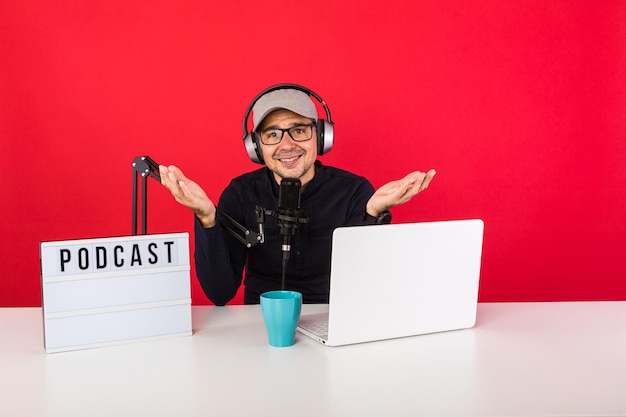 Presentador de hombre con gorra en estudio de grabación de radio podcast haciendo gestos con las manos, junto a una computadora, un micrófono y una caja de luz con la palabra podcast, sobre fondo rojo. Podcasting, concepto de transmisión