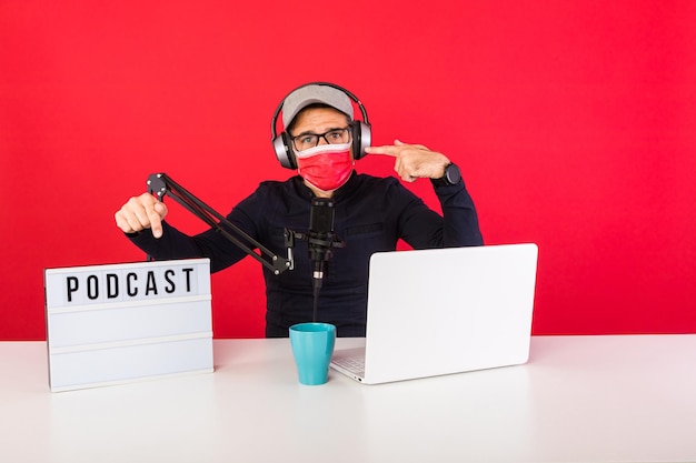 Presentador hombre vestido con gorra roja y máscara en estudio de grabación de radio de podcast, junto a una computadora, un micrófono y una caja de luz con la palabra podcast, apuntando a la máscara, sobre un fondo rojo.