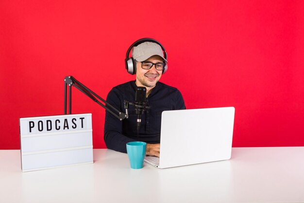 Presentador masculino con gorra en estudio de grabación de radio podcast sonriendo y consultando Internet, junto a una computadora, un micrófono y una caja de luz con la palabra podcast, sobre un fondo rojo.