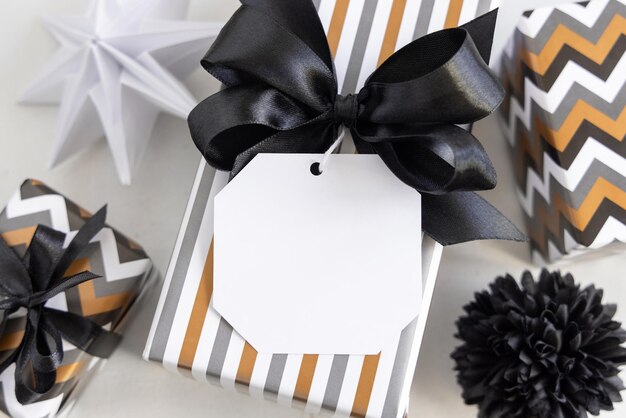 Presentar con etiqueta de regalo en blanco y lazo negro de cerca Cajas de regalo envueltas en papel geométrico rayado y chevron cerca de decoración en blanco y negro Maqueta de etiqueta de aniversario de cumpleaños de año nuevo de Navidad