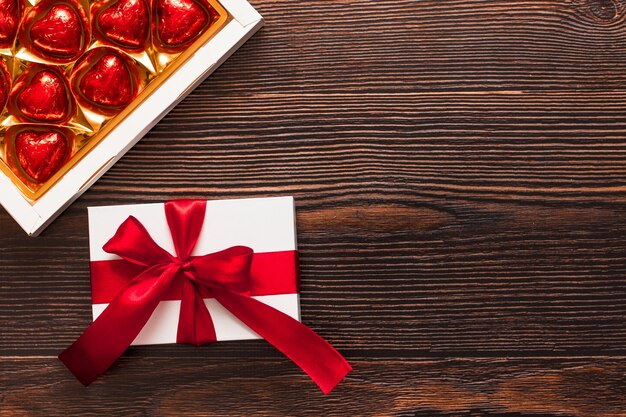 Presente branco com uma fita vermelha e uma caixa de corações de chocolate vermelho isoladas em um fundo escuro de madeira. vista lateral superior de um flatlay quente comemorativo. dia dos namorados e conceito de natal. copyspace.