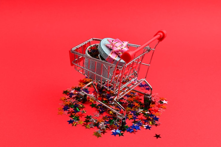Foto presente cuadro azul con lazo rosa en un carrito de compras y confeti sobre un fondo rojo.