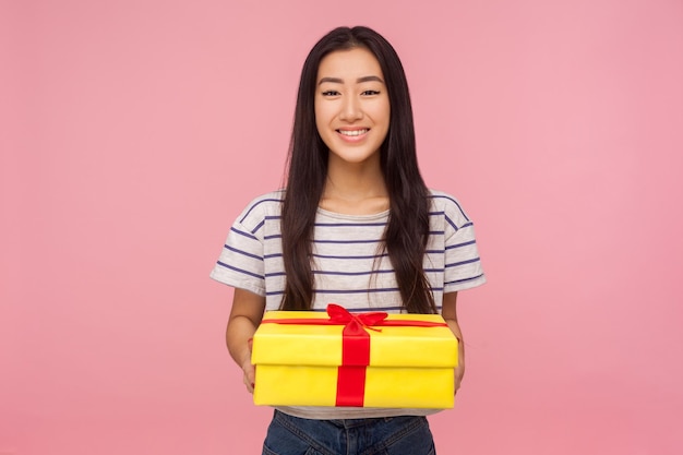 Presente de aniversário. Retrato de menina bonita feliz e satisfeita com cabelo moreno em camiseta listrada, segurando a caixa de presente e sorrindo para a câmera, comemorando feriados. tiro de estúdio isolado no fundo rosa