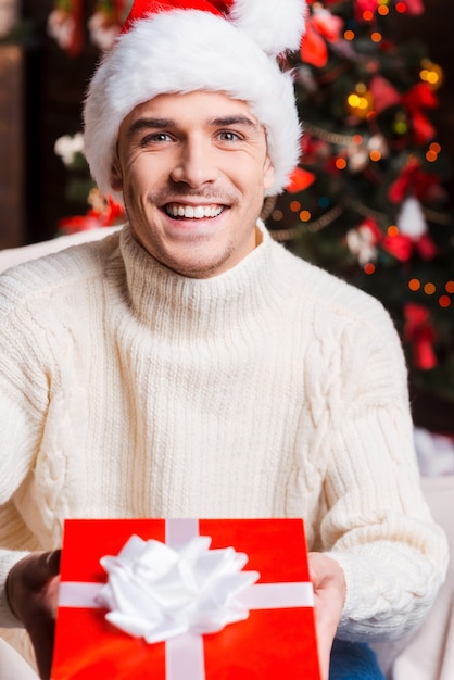 Presente de Natal para você! Jovem bonito com chapéu de Papai Noel estendendo uma caixa de presente e sorrindo com uma árvore de Natal ao fundo