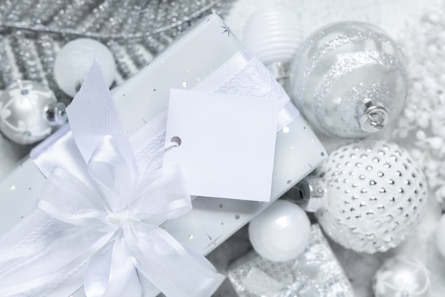 Presente embrulhado com um laço branco e etiqueta quadrada de papel em uma mesa branca com vista superior das decorações de Natal brancas e prateadas. Composição de inverno com cartão de etiqueta em branco, maquete, espaço de cópia