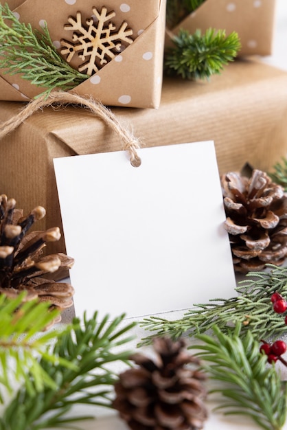 Presente embrulhado de Natal com etiqueta de presente de papel quadrado em uma mesa branca com galhos de árvores de abeto e decorações close-up. Composição rústica de inverno com maquete de etiqueta de presente em branco, espaço de cópia
