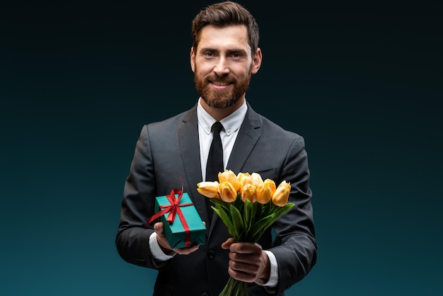 Presentes de aniversário. Encantador prazer romântico jovem em terno formal, segurando o buquê de flores amarelas e um presente com laço vermelho e sorriso dentuço. Interior, foto de estúdio, isolado em fundo azul