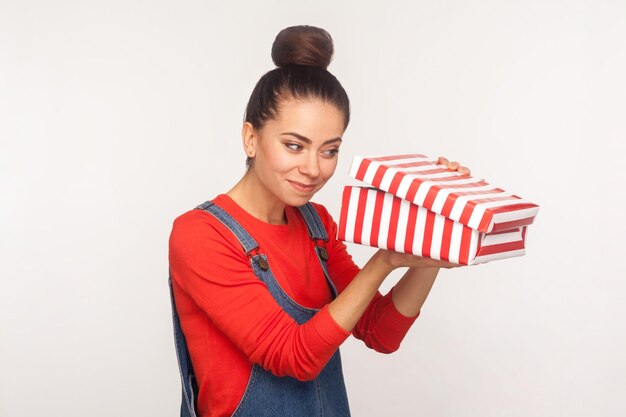 Presentes de feriado. retrato de menina bonita intrometida feliz com coque de cabelo em macacão jeans sorrindo e olhando para a caixa de presente com curiosa expressão de espera, imaginando o que há dentro. tiro de estúdio, isolado