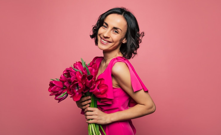 Foto presentes de março. foto de close-up de uma mulher deslumbrante em uma roupa rosa brilhante, que está olhando para a câmera com um largo sorriso, segurando um ramo de flores da primavera.