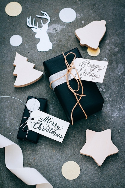 Presentes de Natal em embalagem preta com etiquetas, confetes e decoração em madeira sobre um fundo cinza